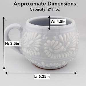 Op de afbeelding: Een lichtgrijze keramische mok met een wit reli&euml;f bloemenpatroon. De mok heeft een inhoud van 621 ml, een hoogte van 8,89 cm, een breedte van 11,43 cm en een lengte van 15,88 cm. De onderkant van de mok toont een roodbruine kleikleur.