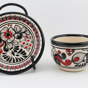 May include: A white ceramic teacup and saucer set with black and red floral designs. The cup has a handle and the saucer has a black rim.