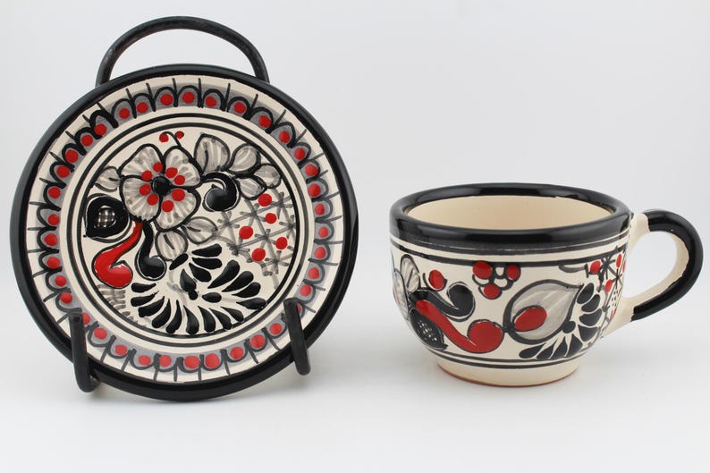 May include: A white ceramic teacup and saucer set with black and red floral designs. The cup has a handle and the saucer has a black rim.