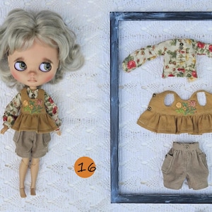 Puede incluir: Una mu&ntilde;eca de cabello claro ondulado viste una camisa de manga larga con flores, un chaleco bordado color mostaza y pantalones cortos beige. Junto a la mu&ntilde;eca, un marco rectangular contiene una camisa floral, un chaleco y pantalones cortos.