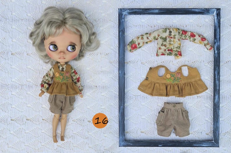 Puede incluir: Una mu&ntilde;eca de cabello claro ondulado viste una camisa de manga larga con flores, un chaleco bordado color mostaza y pantalones cortos beige. Junto a la mu&ntilde;eca, un marco rectangular contiene una camisa floral, un chaleco y pantalones cortos.