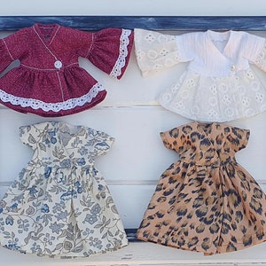 K&ouml;nnte beinhalten: Vier Puppenkleider in einer blauen Rahmendekoration. Die Kleider umfassen ein weinrotes Wickeloberteil mit Glocken&auml;rmeln, ein wei&szlig;es Spitzen-Wickeloberteil, ein Kleid mit Blumenmuster und ein Kleid mit Leopardenmuster. Jedes Kleid hat einen Knopfverschluss.