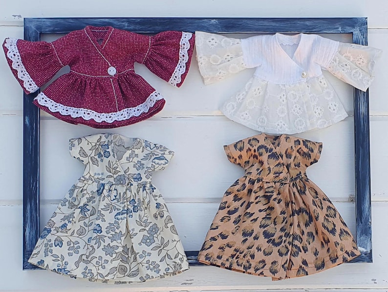 K&ouml;nnte beinhalten: Vier Puppenkleider in einer blauen Rahmendekoration. Die Kleider umfassen ein weinrotes Wickeloberteil mit Glocken&auml;rmeln, ein wei&szlig;es Spitzen-Wickeloberteil, ein Kleid mit Blumenmuster und ein Kleid mit Leopardenmuster. Jedes Kleid hat einen Knopfverschluss.