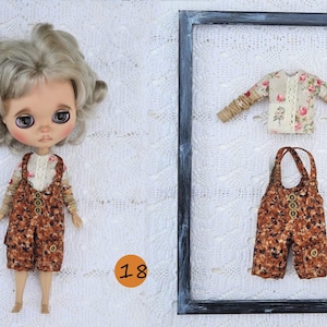 Puede incluir: Una mu&ntilde;eca de pelo rubio ondulado viste una camisa de flores de manga larga y un peto marr&oacute;n estampado con botones. Junto a la mu&ntilde;eca hay un marco negro que contiene una camisa de flores y un peto a juego con botones.