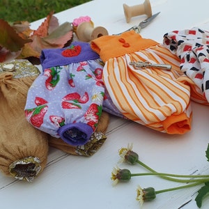 May include: Four handmade doll dresses in different patterns and colors. The dresses are made of cotton fabric and have a variety of details, including ruffles, buttons, and embroidery. The dresses are displayed on a white surface.