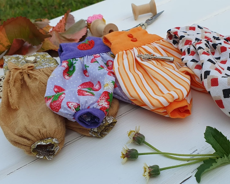 May include: Four handmade doll dresses in different patterns and colors. The dresses are made of cotton fabric and have a variety of details, including ruffles, buttons, and embroidery. The dresses are displayed on a white surface.