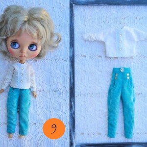 Puede incluir: Una mu&ntilde;eca con cabello rubio ondulado y grandes ojos azules que viste una blusa blanca de manga larga con tres botones y pantalones turquesa. Al lado hay una blusa blanca a juego y pantalones turquesa con un bot&oacute;n y detalles dorados.