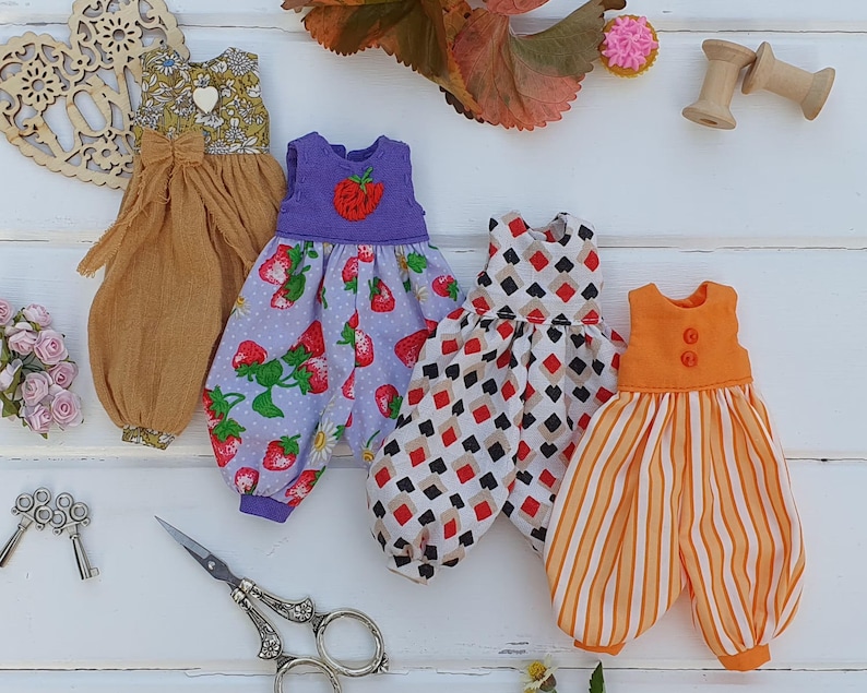 May include: Four handmade doll dresses in different patterns and colors. The dresses are made of cotton fabric and have a variety of details, including ruffles, buttons, and embroidery.