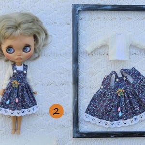 Puede incluir: Una mu&ntilde;eca de pelo rubio viste una camiseta blanca de manga larga y un vestido de tirantes azul oscuro con estampado floral y encaje blanco. Al lado, un conjunto a juego compuesto por una camiseta blanca y un vestido en un marco.