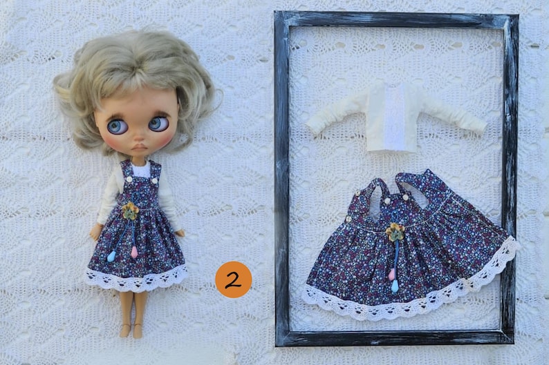 Puede incluir: Una mu&ntilde;eca de pelo rubio viste una camiseta blanca de manga larga y un vestido de tirantes azul oscuro con estampado floral y encaje blanco. Al lado, un conjunto a juego compuesto por una camiseta blanca y un vestido en un marco.