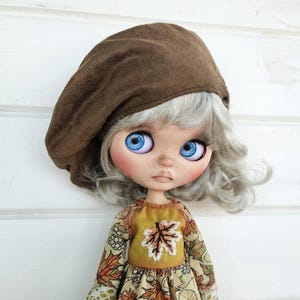 May include: A doll with grey hair wearing a brown beret and a yellow and brown dress with a leaf design. The doll has blue eyes and a neutral expression.