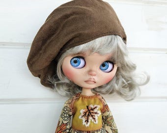 May include: A doll with grey hair wearing a brown beret and a yellow and brown dress with a leaf design. The doll has blue eyes and a neutral expression.