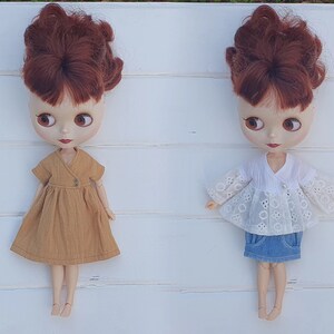 K&ouml;nnte beinhalten: Zwei Blythe-Puppen mit kastanienbraunem Haar und gro&szlig;en Augen. Eine Puppe tr&auml;gt ein senffarbenes Kleid mit Wickeloberteil. Die andere Puppe ist mit einem wei&szlig;en Lochspitzentop und einem Jeansrock bekleidet. Beide Puppen stehen auf einer wei&szlig;en Oberfl&auml;che.