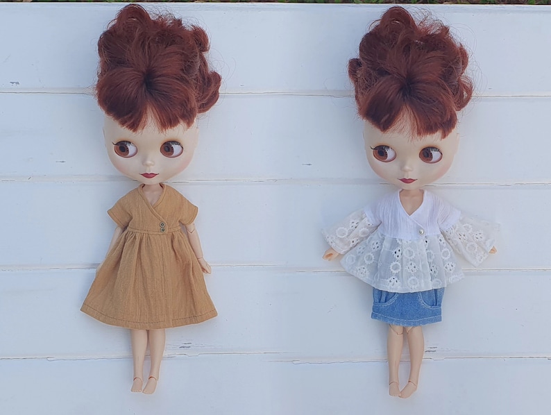 K&ouml;nnte beinhalten: Zwei Blythe-Puppen mit kastanienbraunem Haar und gro&szlig;en Augen. Eine Puppe tr&auml;gt ein senffarbenes Kleid mit Wickeloberteil. Die andere Puppe ist mit einem wei&szlig;en Lochspitzentop und einem Jeansrock bekleidet. Beide Puppen stehen auf einer wei&szlig;en Oberfl&auml;che.