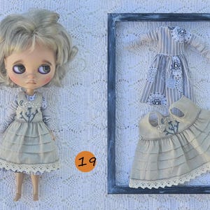 Puede incluir: Una mu&ntilde;eca de cabello rubio ondulado viste un vestido beige con falda de capas y bordado floral, junto a una camisa de rayas de manga larga. A su lado, un vestido a juego y una blusa de rayas de manga larga se exhiben dentro de un marco oscuro.