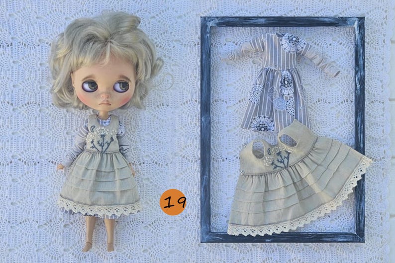 Puede incluir: Una mu&ntilde;eca de cabello rubio ondulado viste un vestido beige con falda de capas y bordado floral, junto a una camisa de rayas de manga larga. A su lado, un vestido a juego y una blusa de rayas de manga larga se exhiben dentro de un marco oscuro.