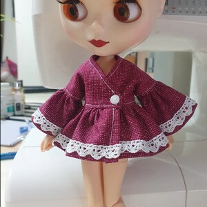 K&ouml;nnte beinhalten: Eine Puppe in einem bordeauxfarbenen Kleid mit wei&szlig;er Spitzenborte und Glocken&auml;rmeln. Das Kleid hat einen wei&szlig;en Knopfverschluss und einen V-Ausschnitt. Die Puppe hat gro&szlig;e braune Augen und roten Lippenstift.