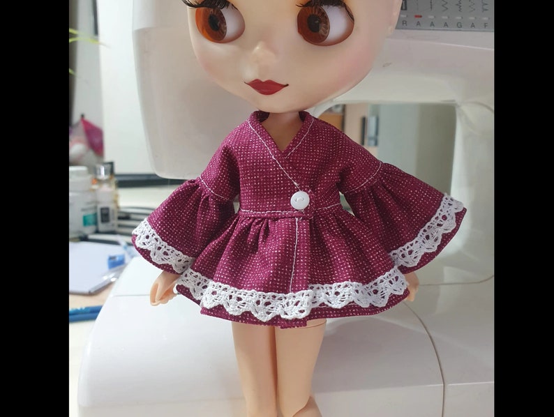 K&ouml;nnte beinhalten: Eine Puppe in einem bordeauxfarbenen Kleid mit wei&szlig;er Spitzenborte und Glocken&auml;rmeln. Das Kleid hat einen wei&szlig;en Knopfverschluss und einen V-Ausschnitt. Die Puppe hat gro&szlig;e braune Augen und roten Lippenstift.