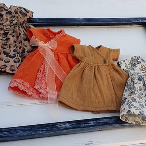 K&ouml;nnte beinhalten: Vier Miniaturkleider in verschiedenen Mustern und Farben werden gezeigt. Eines hat einen Leopardenmuster, ein anderes ist orange mit Spitzenbesatz und einer Schleife, ein drittes ist senffarben und das letzte ist floral mit blauen Akzenten. Sie sind in einem dunkelblauen Rahmen angeordnet.