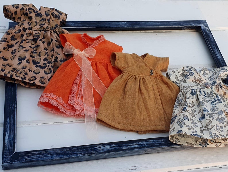 K&ouml;nnte beinhalten: Vier Miniaturkleider in verschiedenen Mustern und Farben werden gezeigt. Eines hat einen Leopardenmuster, ein anderes ist orange mit Spitzenbesatz und einer Schleife, ein drittes ist senffarben und das letzte ist floral mit blauen Akzenten. Sie sind in einem dunkelblauen Rahmen angeordnet.