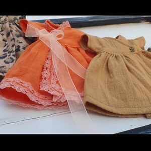 K&ouml;nnte beinhalten: Eine Sammlung von Miniaturkleidern in verschiedenen Farben und Mustern. Ein orangefarbenes Kleid mit Spitzenbesatz und einer durchsichtigen Schleife steht im Vordergrund. Weitere Kleider sind ein beigefarbenes Kleid mit Kn&ouml;pfen und ein Leopardenmusterkleid.