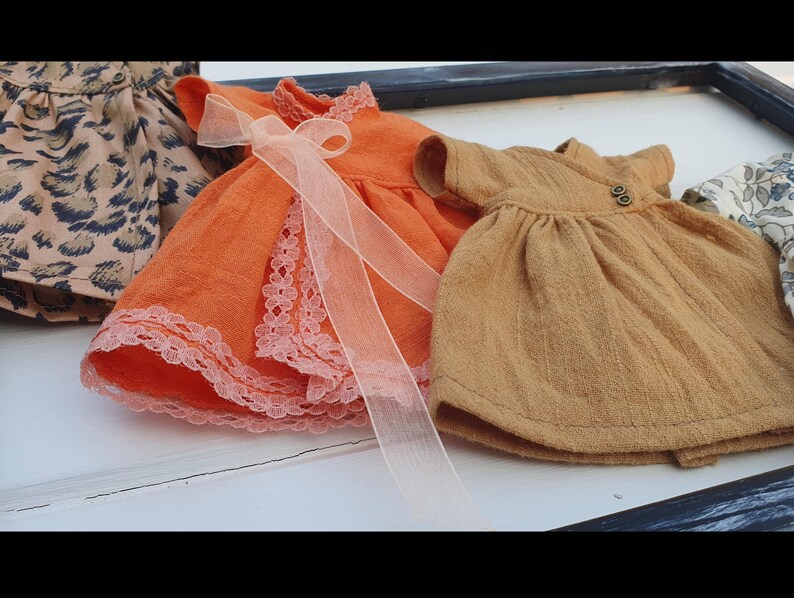 K&ouml;nnte beinhalten: Eine Sammlung von Miniaturkleidern in verschiedenen Farben und Mustern. Ein orangefarbenes Kleid mit Spitzenbesatz und einer durchsichtigen Schleife steht im Vordergrund. Weitere Kleider sind ein beigefarbenes Kleid mit Kn&ouml;pfen und ein Leopardenmusterkleid.