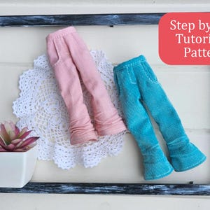 Op de afbeelding: Twee paar poppenbroeken, een roze en een turquoise, tentoongesteld in een zwart frame met een witte kant en een kleine plant in een pot. De tekst "Stap voor stap tutorial & patroon" wordt weergegeven in een rode doos.