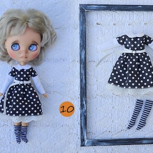 Puede incluir: Una mu&ntilde;eca de cabello rubio ondulado y ojos azules viste un vestido negro con lunares blancos, cuello blanco y mangas largas blancas, junto con calcetines a rayas. Un vestido y calcetines a rayas a juego se exhiben dentro de un marco negro.