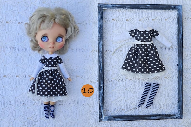 Puede incluir: Una mu&ntilde;eca de cabello rubio ondulado y ojos azules viste un vestido negro con lunares blancos, cuello blanco y mangas largas blancas, junto con calcetines a rayas. Un vestido y calcetines a rayas a juego se exhiben dentro de un marco negro.