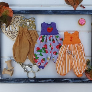 May include: Three handmade doll outfits in a black frame. The outfits are made of different fabrics and have different patterns. The first outfit is a brown and white floral print with a brown sash. The second outfit is a purple and white floral print with a strawberry design. The third outfit is a striped orange and white outfit with a small key charm.