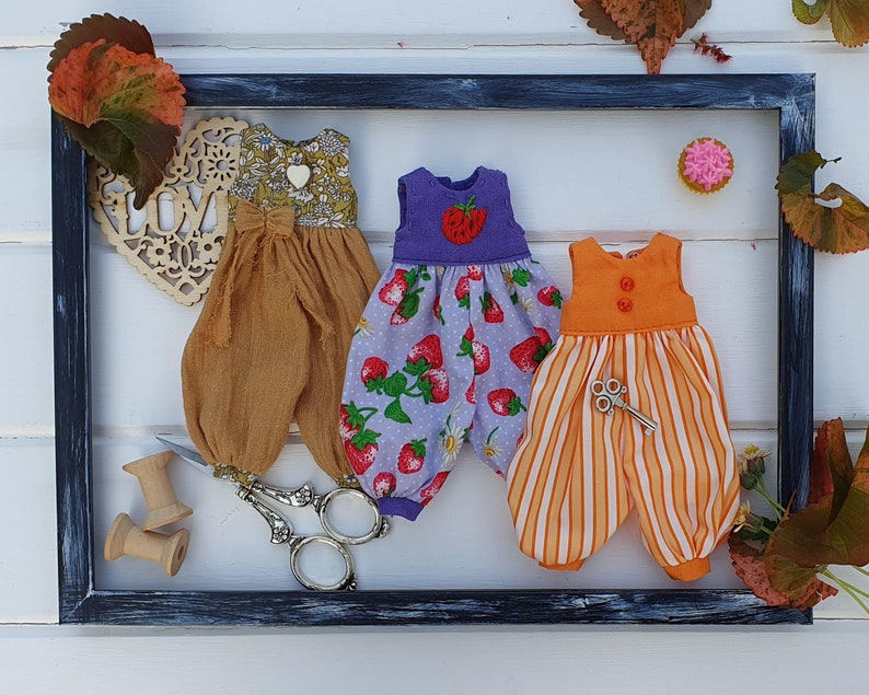 May include: Three handmade doll outfits in a black frame. The outfits are made of different fabrics and have different patterns. The first outfit is a brown and white floral print with a brown sash. The second outfit is a purple and white floral print with a strawberry design. The third outfit is a striped orange and white outfit with a small key charm.