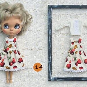 Puede incluir: Una mu&ntilde;eca con cabello rubio ondulado viste una camisa blanca de manga larga y un vestido tipo pichi color crema con estampado de fresas rojas, botones rojos y encaje blanco. Al lado, un conjunto a juego con camisa y vestido.