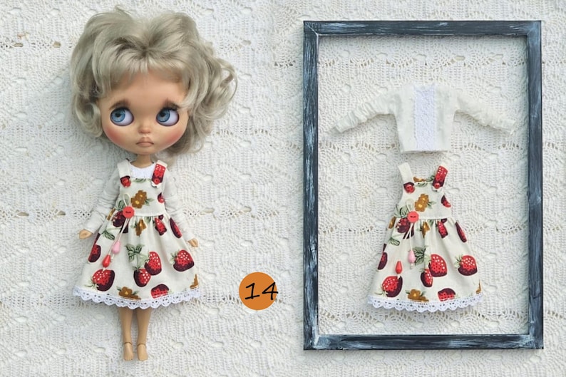 Puede incluir: Una mu&ntilde;eca con cabello rubio ondulado viste una camisa blanca de manga larga y un vestido tipo pichi color crema con estampado de fresas rojas, botones rojos y encaje blanco. Al lado, un conjunto a juego con camisa y vestido.