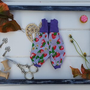 May include: A purple and white fabric doll outfit with a strawberry print. The outfit is displayed in a black frame with a wooden heart, leaves, and a small pink cupcake.