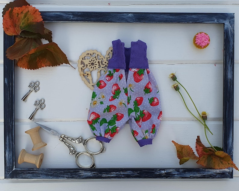 May include: A purple and white fabric doll outfit with a strawberry print. The outfit is displayed in a black frame with a wooden heart, leaves, and a small pink cupcake.