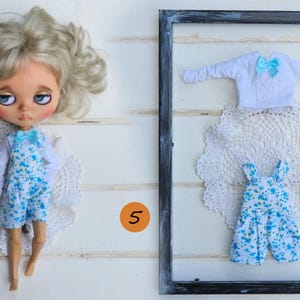 Puede incluir: Una mu&ntilde;eca de cabello rubio viste una camisa blanca de manga larga y un overol azul con estampado floral y un lazo azul claro. Al lado, un conjunto enmarcado incluye una camisa blanca de manga larga con un lazo azul claro y un overol floral a juego.