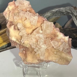 May include: A natural mineral specimen featuring a cluster of translucent, cubic crystals atop a matrix of peach and cream-colored stone. The mineral is displayed on a clear acrylic stand, showcasing its unique texture and form.