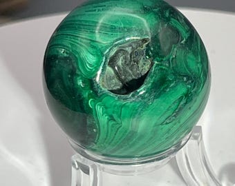 May include: A green malachite sphere with a natural hole, displayed on a clear acrylic stand.