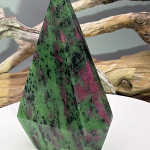 May include: A faceted, pyramid-shaped stone with a green base color, accented with streaks of magenta and black. The gemstone is polished and has a crystalline appearance. The object is displayed on a white surface, with driftwood in the background.