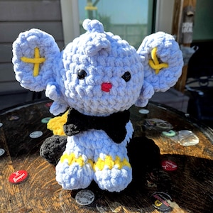 Puede incluir: Un peluche de elefante de ganchillo azul claro hecho a mano con orejas grandes, detalles de cruz amarillos y una pajarita negra. El elefante tiene ojos negros y una nariz roja. El juguete está sobre una superficie de madera.
