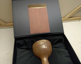 May include: A matte brown chalice with gold leaf accents rests inside a black presentation box. The box lid features a rectangular brown envelope with handwritten text. The chalice is nestled in black fabric lining within the box interior.