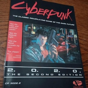 以下が含まれることがあります： 黒色のサイバーパンク2020セカンドエディションのロールプレイングゲームの表紙。表紙には、銃を持った男性の未来的なイラストが描かれており、ゲームのタイトルは赤色です。「The Classic Roleplaying Game of the Dark Future」というテキストと引用が含まれています。
