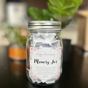 May include: A clear glass jar with a silver lid filled with folded pieces of paper. The jar has a pink and white floral label that says "Happy Anniversary Memory Jar" and includes the text "Thanks for all of our greatly shared moments. This year has been full of love and joy. Let's fill our next year with even more memorable moments and capture them all in this jar! Love you."