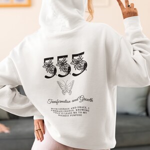 May include: White hooded sweatshirt with a black graphic print of the number 555 with flowers and a butterfly. The text "Transformation and Growth" is printed below the number with the text "With courage and grace, I embrace change, knowing that it leads me to my highest purpose."