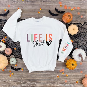 May include: White sweatshirt with the text "Life is Short" in colorful letters and a black heart. The left sleeve has the text "Make it Sweet" in colorful letters.