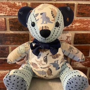 May include: A blue and white patchwork teddy bear with a dinosaur print. The bear has a blue bow tie and black button eyes.