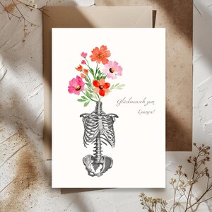 Könnte beinhalten: Grußkarte mit einer Aquarellillustration eines menschlichen Skelettorso mit einem Strauß roter und rosa Blumen. Auf der Karte steht der Text "Glückwunsch zum Examen!" in Schrift.