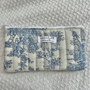 Puede incluir: Un neceser acolchado con un estampado de escena pastoral en azul y blanco. La bolsa tiene cierre de cremallera y una pequeña etiqueta que dice "scarlett creates". La bolsa es rectangular y tiene líneas acolchadas verticales.