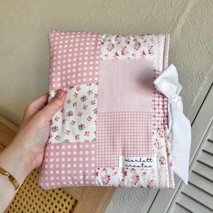 Könnte beinhalten: Handgefertigte Buchhülle im Patchwork-Stil. Sie zeigt eine Mischung aus rosa Vichy-, Blumen- und Streifenstoffen. Eine weiße Schleife hält die Hülle zusammen, und ein kleines Etikett trägt die Aufschrift "scarlett creates".