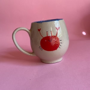 Könnte beinhalten: Ein runder, cremeweißer Keramikbecher mit einer handgemalten roten Krabbenillustration auf der Vorderseite. Die Krabbe hat zwei erhobene Scheren, dünne schwarze Augen und kleine schwarze Beine. Der Becher hat einen geschwungenen Henkel und einen blauen Rand.